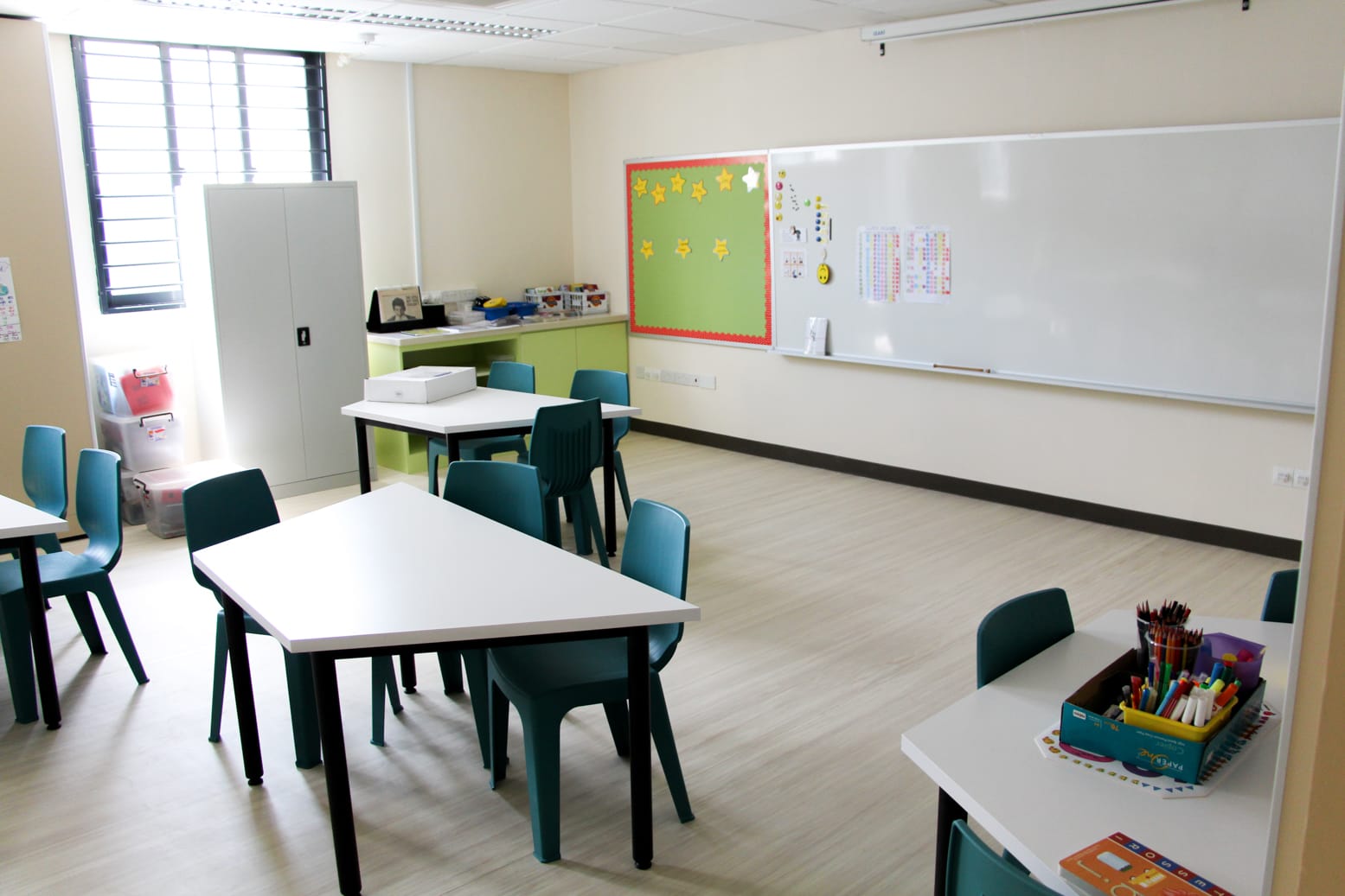 Empty classroom with teal chairs, white tables, a whiteboard, and a green bulletin board with yellow stars.